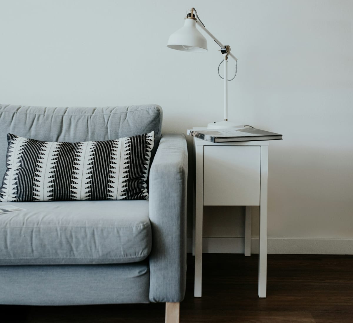 a photo of half of a grey couch on a wooden floor. A small white table with a white lamp and a book on top, is beside the couch.