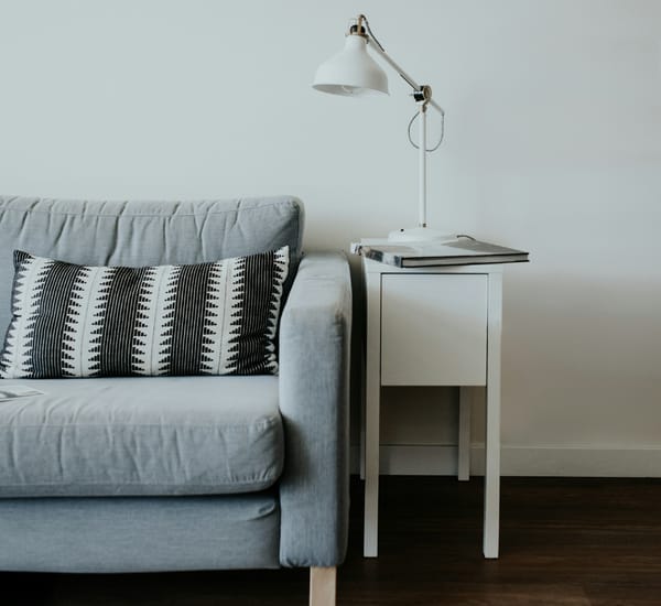 a photo of half of a grey couch on a wooden floor. A small white table with a white lamp and a book on top, is beside the couch.