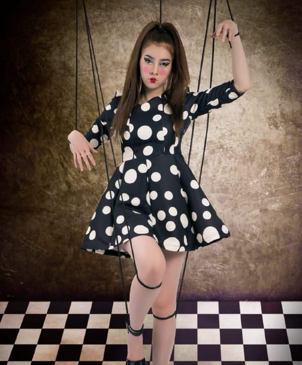 A woman is dressed like a doll, and puppet strings are attached to her limbs. Shes standing on a chessboard.